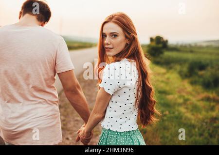 Rückansicht eines Paares, das die Hände hält und auf einer Landstraße läuft. Mädchen mit roten Haaren und Sommersprossen, die ihren Kopf zur Kamera drehen. Sommer Stockfoto