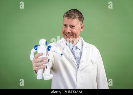 Wissenschaftler mit weißem Mantel hält einen kleinen weißen Roboter in der Hand und steht vor grünem Hintergrund Stockfoto