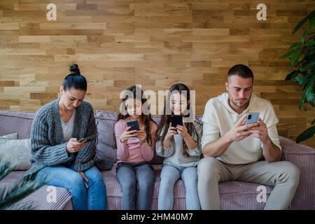 Familie mit zwei kleinen Töchtern, jeder nutzt Handy im Wohnzimmer. Stockfoto