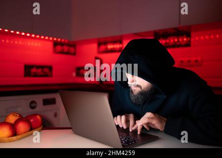 Ein Hacker in einem schwarzen Hoodie mit einer Kapuze auf dem Kopf sitzt an einem Laptop, der sich über das Internet gebeugt hat Stockfoto