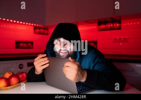 Ein Hacker in einem schwarzen Hoodie mit einer Kapuze auf dem Kopf sitzt an einem Laptop und greift mit seinen Händen an einen Laptop Stockfoto