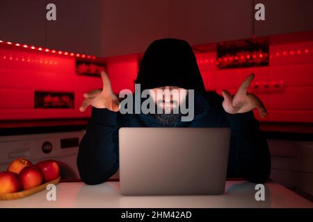 Ein Hacker in einem schwarzen Hoodie mit einer Kapuze auf dem Kopf sitzt an einem Laptop und zeigt seine Daumen nach oben und freut sich über einen erfolgreichen Hack Stockfoto