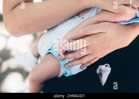Unterer Teil eines Säuglings, der von einer nicht erkennbaren Frau in ihren Armen gehalten wird. Selektiver Fokus. Stockfoto
