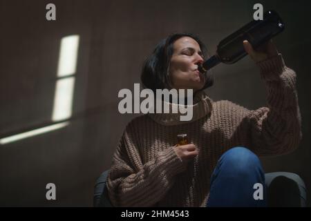 Depressive betrunkene Frau, die im Dunkeln auf dem Boden sitzt und Pillen nimmt und Wein aus einer Flasche trinkt Stockfoto