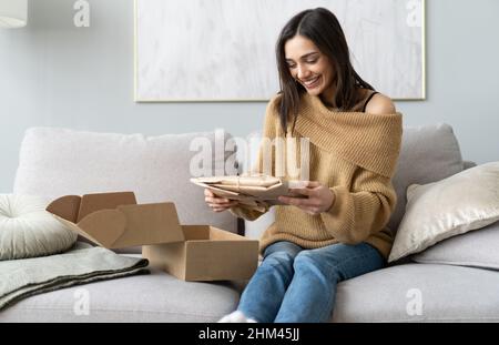 Lächelnd glücklich junge Frau sitzt auf der Couch im Wohnzimmer, Auspacken Karton Paket online bestellen aus dem internationalen Internet-Shop, zufrieden mit Stockfoto