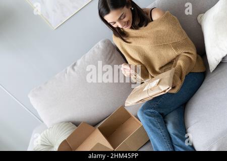 Lächelnd glücklich junge Frau sitzt auf der Couch im Wohnzimmer, Auspacken Karton Paket online bestellen aus dem internationalen Internet-Shop, zufrieden mit Stockfoto