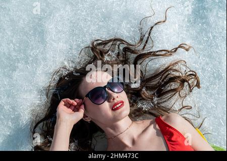 Nahaufnahme einer kaukasischen Frau, die in einer Sonnenbrille auf dem Schnee liegt. Draufsicht. Stockfoto