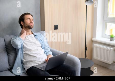 Porträt eines müden, tausendjährigen jungen Mannes, Freiberufler oder Student, sitzt mit einem Laptop auf dem Sofa, müder Kerl massiert sich den Nacken, fühlt sich von der Fernarbeit Burnout Stockfoto