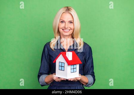 Foto von schönen blonde reife Dame tragen Jeans Kleid halten Haus isoliert auf grünem Hintergrund Stockfoto