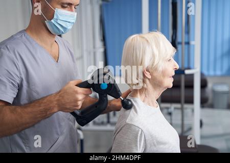 Physiotherapeut behandelt die Schulter des Rentners mit einer Muskelmassagepistole Stockfoto