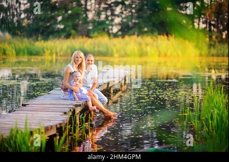 Glückliche Familie, die sich auf einem hölzernen Pier ausruhte, ihre Füße ins Wasser tauche, Zeit für die Familie Stockfoto