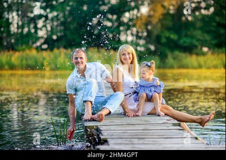 Glückliche Familie, die sich auf einem hölzernen Pier ausruhen, ihre Füße ins Wasser tauchen und Spray machen, Familienzeit Stockfoto