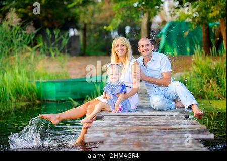 Glückliche Familie, die sich auf einem hölzernen Pier ausruhen, ihre Füße ins Wasser tauchen und Spray machen, Familienzeit Stockfoto