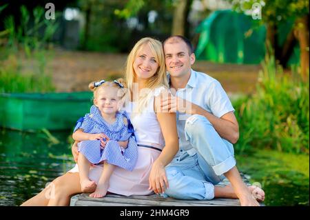 Glückliche Familie, die sich auf einem hölzernen Pier ausruhte, ihre Füße ins Wasser tauche und lächle, Zeit für die Familie Stockfoto