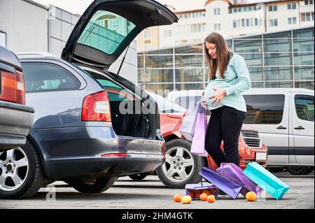 Eine Schwangerin mit Papiertüten gebärt ein frühes Kind auf dem Parkplatz in der Innenstadt. Junge Mutter mit Bauch Bauchgefühl Schmerzen während gebrochenem Wasser nach dem Einkaufen während der Erwartungszeit. Stockfoto