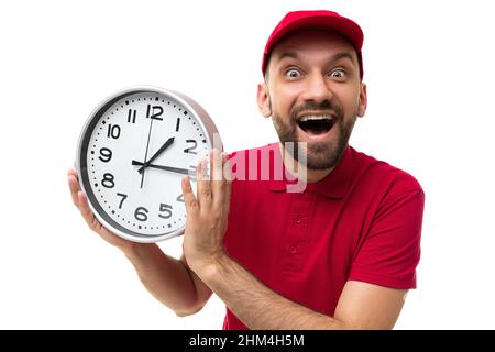 Charismatischer Kurier, der eine Uhr mit einem Lächeln auf dem Gesicht in einem roten T-Shirt und einer Mütze auf weißem Hintergrund hält, das Konzept der schnellen Lieferung und der Zeit Stockfoto