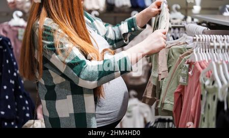 Nahaufnahme des Bauches einer Schwangeren rothaarigen Frau im Kindergeschäft, im Einkaufszentrum, in der Saison der Rabatte und Verkäufe. Werdende Mutter wählt Gerinnsel Stockfoto