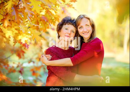 Das süße Mädchen umarmt ihre ältere Mutter, die in einem wunderschönen Herbstpark steht, und umarmt und lächelt auf dem Hintergrund des hellen Laubs, Generationen Stockfoto