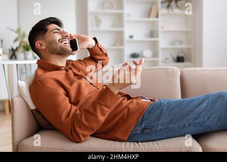 Ein fröhlicher Mann, der auf dem Handy spricht und auf der Couch sitzt Stockfoto