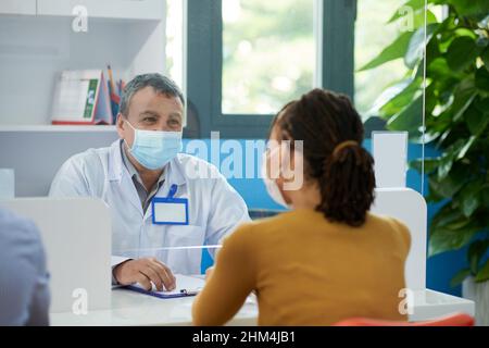 Leitender Hausarzt gibt Empfehlungen an Patienten, wenn sie aufgrund einer Coronavirus-Pandemie mit einer Glasscheibe zwischen ihnen sitzen Stockfoto