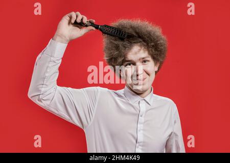 Ein lockiger Mann in einem Hemd lächelt und bürstet sein lockiges Haar. Kämmen Sie lockiges Haar. Salon. Stockfoto