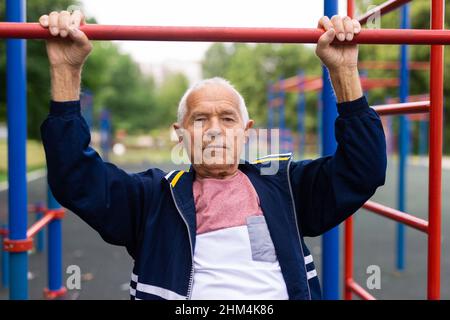 Der alte Mann zieht sich auf dem Sportplatz an der horizontalen Stange hoch. Outdoor-Sport im Alter Stockfoto