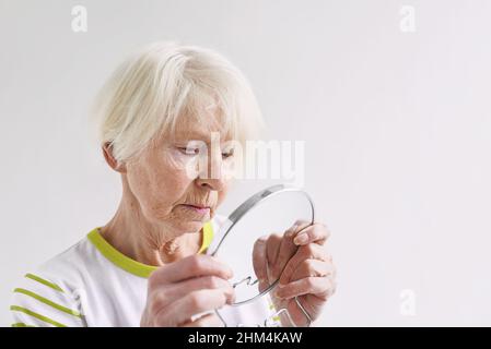 Ältere traurige Frau, die sich im Spiegel ansieht. Alter, Zeit, Hautpflegekonzept Stockfoto