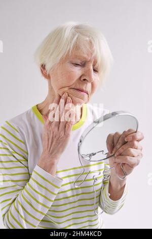 Ältere traurige Frau, die sich im Spiegel ansah und ihr Gesicht berührte. Alter, Zeit, Hautpflegekonzept Stockfoto