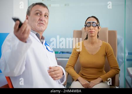 Augenärztin bittet die Patientin in den Testframes, Briefe auf der an der Wand hängenden Testkarte zu lesen, wenn sie ihr Sehvermögen überprüft Stockfoto