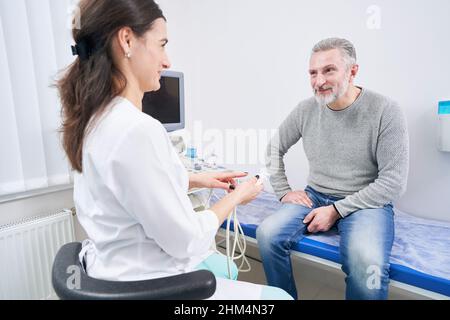 Freundlicher junger Ultraschallforscher, der vor der Ultraschalluntersuchung mit dem Patienten kommuniziert Stockfoto