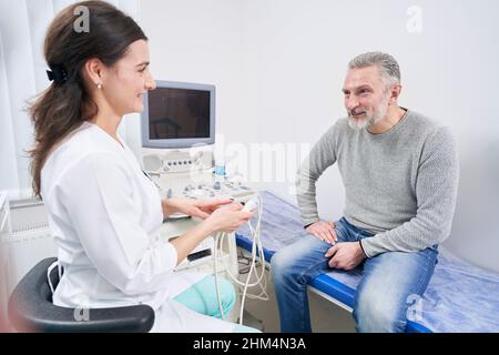 Lächelnder, liebenswürdiger Arzt, der vor der Sonographie mit einem männlichen Patienten spricht Stockfoto