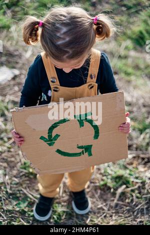 Mädchen mit zwei Zöpfen, die ein Pappschild mit einem Recycling-Symbol halten. Stockfoto