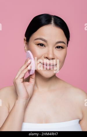 Lächelnde asiatische Frau in Top-Reinigung Gesicht mit Schwamm und Blick auf die Kamera isoliert auf rosa Stockfoto