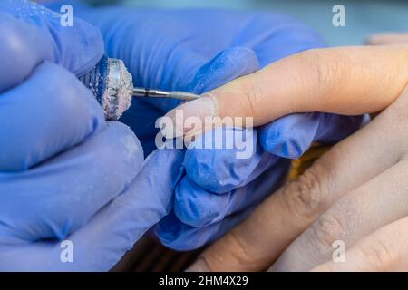 Nahaufnahme der Hände eines Maniküre-Meisters in blauen Handschuhen mit einer elektrischen Nagelfeile zum Trimmen und Entfernen von Nagelhaut in einem Schönheitssalon. Hardware-Maniküre Stockfoto