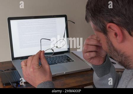 Der Mann arbeitet an einem Laptop und zieht seine Brille aus, weil seine Augen ihn stören. Brille im Vordergrund, Laptop im Hintergrund Stockfoto