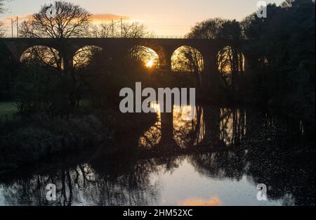 Eisenbahnviadukt bei Sonnenuntergang mit der Sonne, die durch einen der Bögen scheint, was goldenes Leuchten und Reflexionen im Fluss verursacht Stockfoto