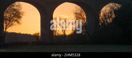 Bögen eines Eisenbahnviadukts in Silhouette mit dem goldenen Licht eines Winters Sonnenuntergang dahinter Stockfoto