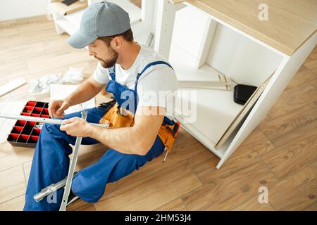Blick aus der Perspektive auf einen konzentrierten Handwerker in Uniform, der auf dem Boden sitzt, während er in der Küche eine Schublade installiert Stockfoto