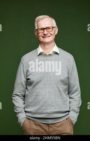 Fröhlicher älterer Mann in Hosen, Pullover und Brillen, der vor der Kamera auf dunkelgrünem Hintergrund steht Stockfoto
