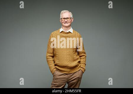 Fröhlicher älterer Mann in Strickpullover und brauner Hose, der vor grauem Hintergrund steht und lächelnd auf die Kamera blickt Stockfoto
