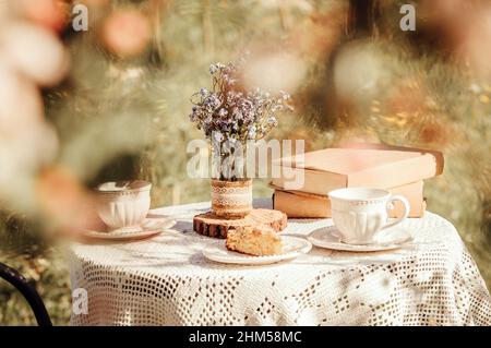 Traumhafter Blick durch Apfelblüten auf den Tisch, mit Teetassen und Büchern. Blumenstrauß Myosotis arvensis Vergiss mich nicht im handgefertigten Glas. Stockfoto