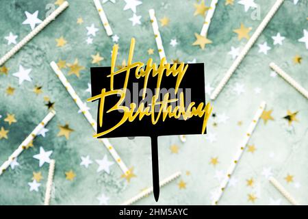 Goldener Schriftzug Happy Birthday auf schwarzem Teller mit Stift auf verschwommenem grünen Hintergrund mit Sternen und Trinkhalmen für Cocktail. Festliche Geburtstagskarte Stockfoto