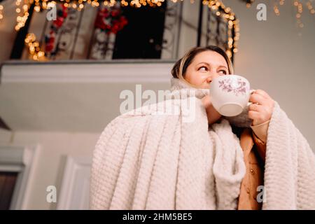 Erwachsene Frau in Winterkleidung mit einer Decke, die draußen vor dem Haus steht und eine Tasse heißen Tee hält und ihn trinkt. Stockfoto