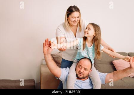 Schöne Familie mit Spaß zu Hause Tochter sitzt auf dem Papa Hals hinter und Vater mit Mutter hält ihre Hände an den Seiten. Stockfoto