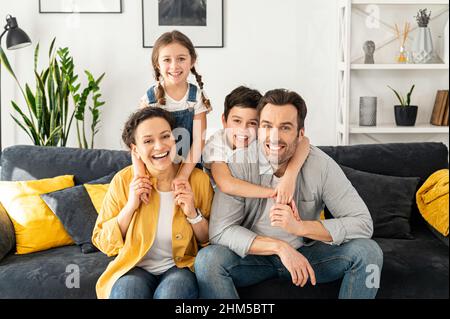 Glücklich zusammen. Porträt einer fröhlichen, multirassischen Familie von vier - Mutter, Vater, Sohn und Kleinkind, die auf dem Sofa im gemütlichen Wohnzimmer sitzen, die Kamera betrachten und lachen, das Wochenende zu Hause genießen Stockfoto