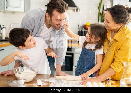 Fröhliche vierköpfige Familie, die zusammen in der Küche kocht, Eltern und zwei Kinder, die zusammen Teig machen, Kuchen oder Kekse zubereiten, am Wochenende Spaß haben, Tochter, die Papa mit Mehl in die Nase schmiert und lacht Stockfoto
