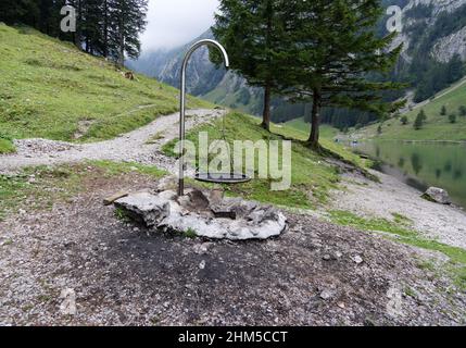 Leerer Kamin mit schwarzem Metallgitter Rost auf großem Kiesplatz, verkohltes Holz zwischen großen Steinen, grüner Tannenwald im Hintergrund, See seealpsee du Stockfoto