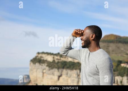 Glücklicher Mann mit schwarzer Haut, der mit der Hand im Berg vor der Sonne schützt und sucht Stockfoto