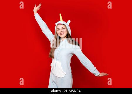 Ein Mädchen im Einhorn-Schlafanzug auf rotem Hintergrund zieht ihre Hand hoch. Übernachtung. Zeit zum Schlafen. Pyjama-Party. Einhorn Kostüm. Ich täuscht mich herum. Stockfoto