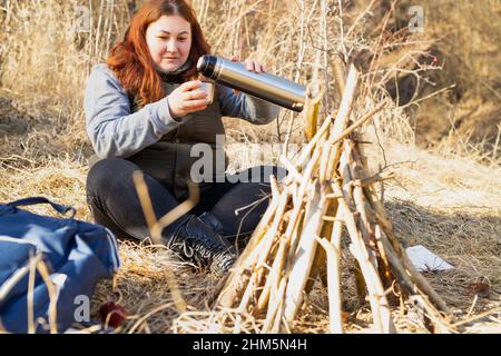 Nahaufnahme einer alleinerziehenden Frau, die sich mit einem Lagerfeuer und einem Rucksack im Wald ausruhte und Tee aus der Thermosflasche goss. Stockfoto